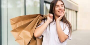 Jovem mulher sorrindo e animada, segurando sacolas de compras em frente a uma loja. A imagem representa o dia de descontos e o entusiasmo com as ofertas da Black Friday.