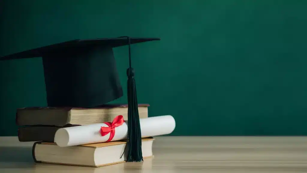 Símbolos da Qualificação: Diploma e Capelo como Prova da Formação Técnica Um diploma enrolado com fita vermelha e um capelo de formatura repousam sobre uma pilha de livros didáticos em uma mesa de madeira.