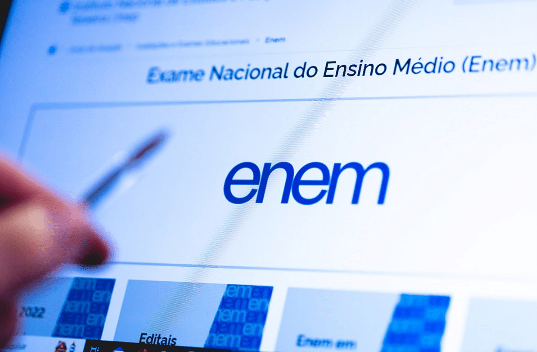 Tela de computador exibindo o site do Exame Nacional do Ensino Médio (Enem) com o logotipo azul "enem" em destaque e uma mão segurando uma caneta apontando para a tela.