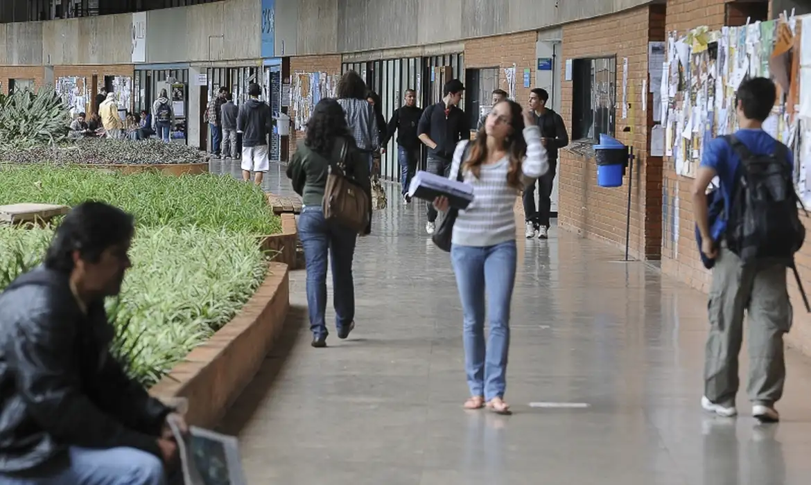 Alunos caminham em corredor de universidade movimentada Pessoas caminhando e interagindo em corredor interno de universidade com murais de avisos