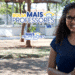 Mulher jovem sorridente com óculos segurando materiais em ambiente escolar ao ar livre.