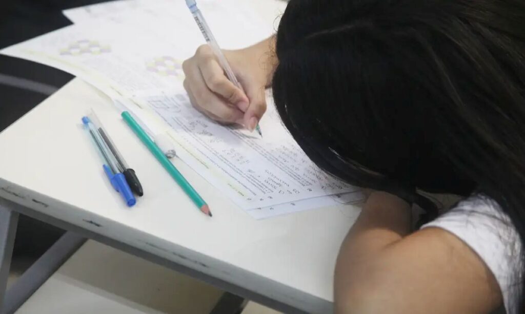 Estudante de cabelo escuro escrevendo ou preenchendo o gabarito de uma prova ou redação com caneta, com cadernos e lápis sobre a mesa, focado na Competência II do ENEM.