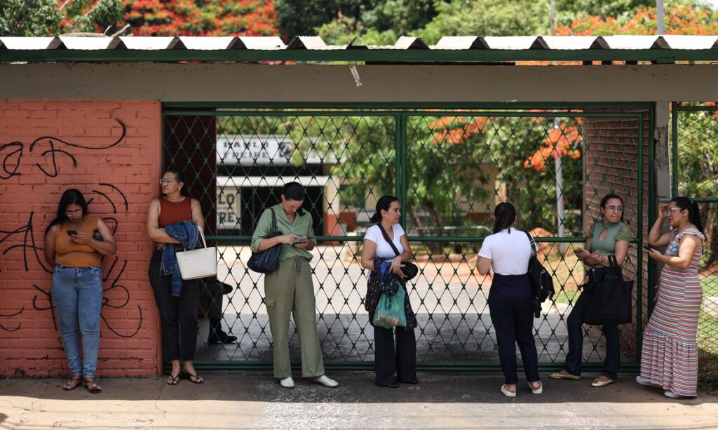 Grupo de candidatos esperando em frente a um portão após a realização da Prova Nacional Docente (PND), verificando informações nos celulares. A cena representa o aguardo dos resultados.
