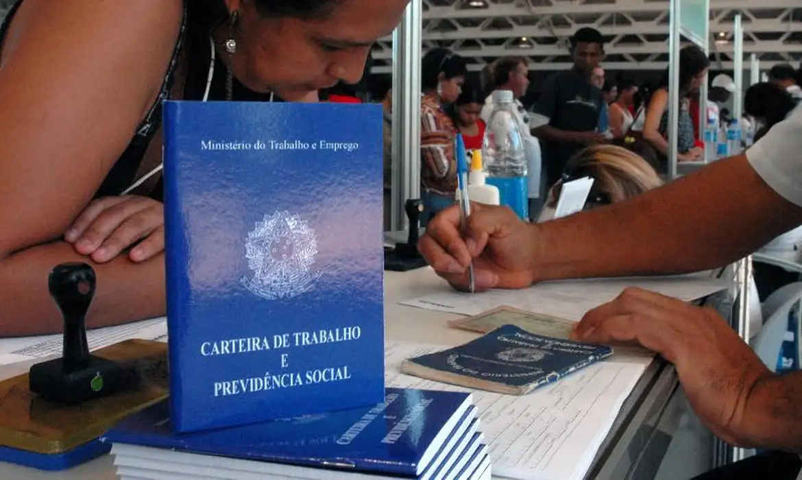 Carteira de Trabalho e Previdência Social em registro Pessoa llenando formulario com varias carteiras de trabalho azuis apiladas sobre escritorio en ambiente de registro laboral
