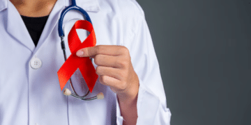 Mão de um médico ou profissional de saúde segurando um laço vermelho, símbolo da campanha Dezembro Vermelho de prevenção e combate ao HIV/AIDS e outras Infecções Sexualmente Transmissíveis (ISTs).