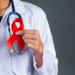 Mão de um médico ou profissional de saúde segurando um laço vermelho, símbolo da campanha Dezembro Vermelho de prevenção e combate ao HIV/AIDS e outras Infecções Sexualmente Transmissíveis (ISTs).