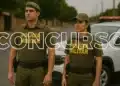 Dois policiais militares de costas com uniforme verde em rua com viatura da polícia ao lado.