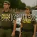 Dois policiais militares de costas com uniforme verde em rua com viatura da polícia ao lado.