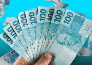 Mão segurando várias cédulas de 100 reais em leque com fundo azul