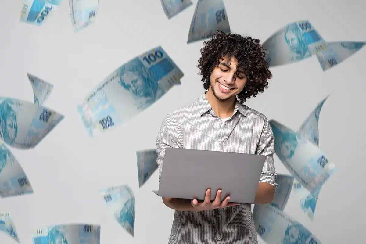 Jovem feliz usando laptop com dinheiro caindo ao redor Homem jovem sorridente segurando notebook com notas de cem reais voando ao redor
