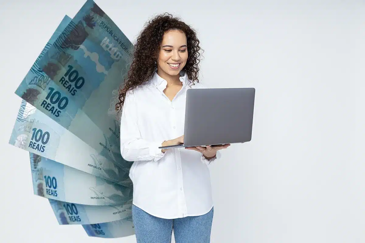 Mulher feliz com laptop e dinheiro em reais ao fundo Mulher sorrindo mexendo em laptop com notas de cem reais em fundo claro