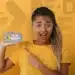 Mulher jovem com blusa amarela apontando para celular com logo do Pé de Meia na tela em fundo amarelo