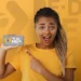 Mulher morena de camiseta amarela segurando celular com logo do Pé de Meia e fundo amarelo