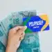 Uma mão segurando um cartão azul e amarelo com a inscrição PIS/PASEP CAIXA, tendo ao fundo várias notas de 100 reais espalhadas em leque.
