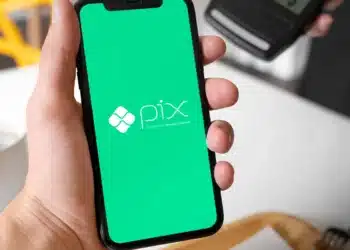 Mão masculina segurando um smartphone que exibe o logotipo branco do Pix em uma tela verde vibrante, com uma máquina de cartões ao fundo.
