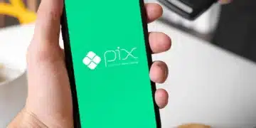 Mão masculina segurando um smartphone que exibe o logotipo branco do Pix em uma tela verde vibrante, com uma máquina de cartões ao fundo.