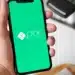 Mão masculina segurando um smartphone que exibe o logotipo branco do Pix em uma tela verde vibrante, com uma máquina de cartões ao fundo.
