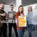 Grupo de quatro profissionais jovens em um escritório moderno, sorrindo e fazendo sinal de positivo, com uma mulher à frente segurando uma prancheta que diz "Vagas de Emprego".