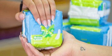 Close das mãos de uma pessoa com unhas feitas e uma tatuagem de coração no pulso, recebendo um pacote de absorventes azuis e verdes em uma farmácia.