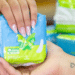 Close das mãos de uma pessoa com unhas feitas e uma tatuagem de coração no pulso, recebendo um pacote de absorventes azuis e verdes em uma farmácia.
