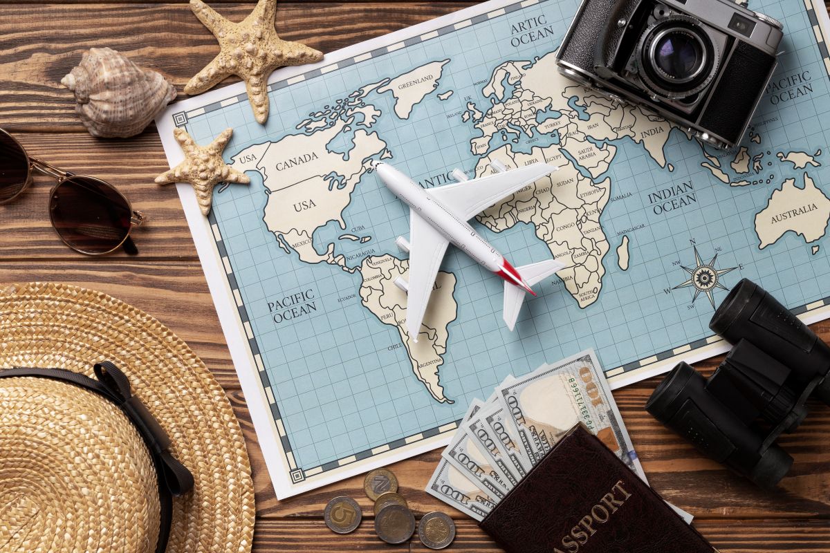 Vista superior de uma mesa de madeira com mapa mútuo, avião de brinquedo, câmera fotográfica retrô, passaporte com notas de dólares, binóculos e chapéu de palha.