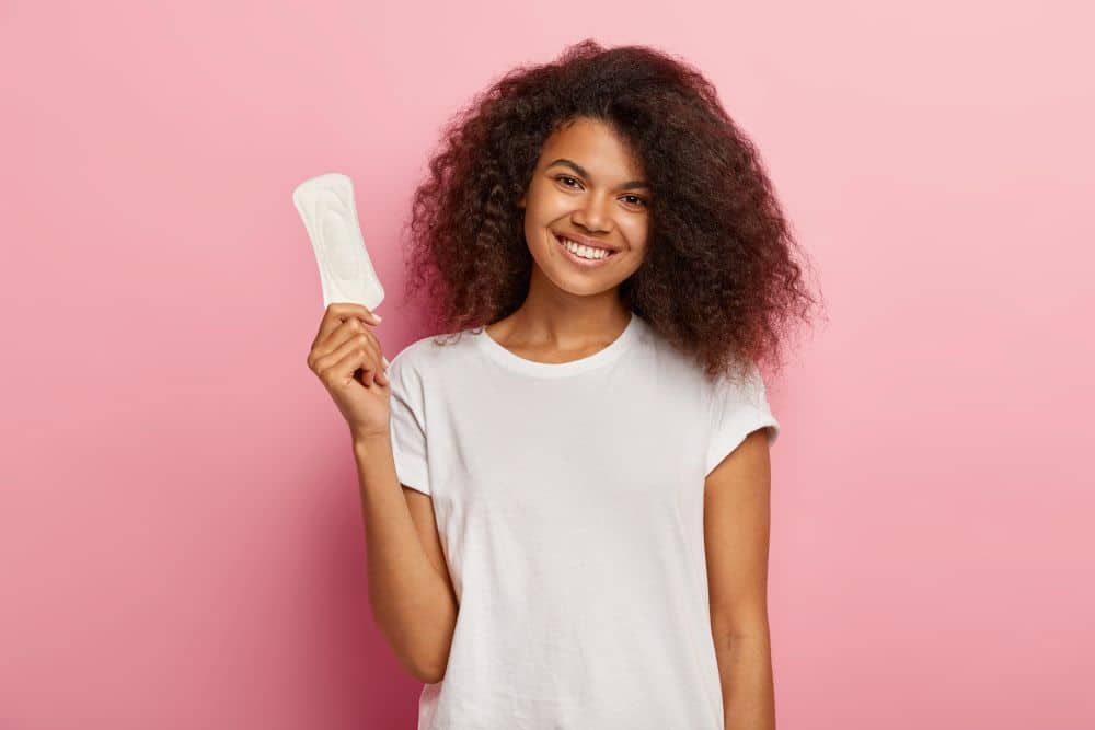 Jovem negra sorridente com cabelos cacheados volumosos, vestindo camiseta branca, segurando um absorvente higiênico descartável contra um fundo rosa.