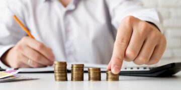 Close de uma mão masculina organizando quatro pilhas crescentes de moedas douradas sobre uma mesa branca, com um homem desfocado ao fundo fazendo anotações.