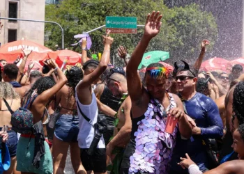 Pessoas celebrando em festa de rua com multidão, sorrisos, e jatos de água sob sol intenso