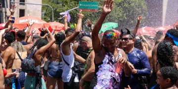Pessoas celebrando em festa de rua com multidão, sorrisos, e jatos de água sob sol intenso