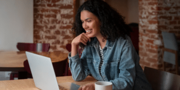 Mulher negra sorridente com jaqueta jeans trabalhando no notebook em uma mesa de madeira com xícara branca