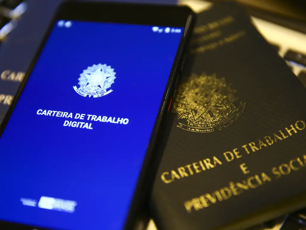 Smartphone com o aplicativo Carteira de Trabalho Digital ao lado da tradicional Carteira de Trabalho e Previdência Social (CTPS) física. A imagem simboliza a transição do documento manual para o formato eletrônico e ágil.