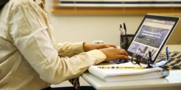 Pessoa usando camisa bege digitando em notebook com caderno, lápis e óculos sobre mesa branca