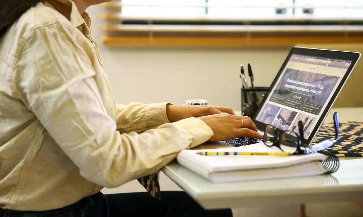 Mulher trabalhando em notebook em ambiente de escritório moderno Pessoa digitando em notebook com roupas claras em mesa com caderno e óculos em escritório
