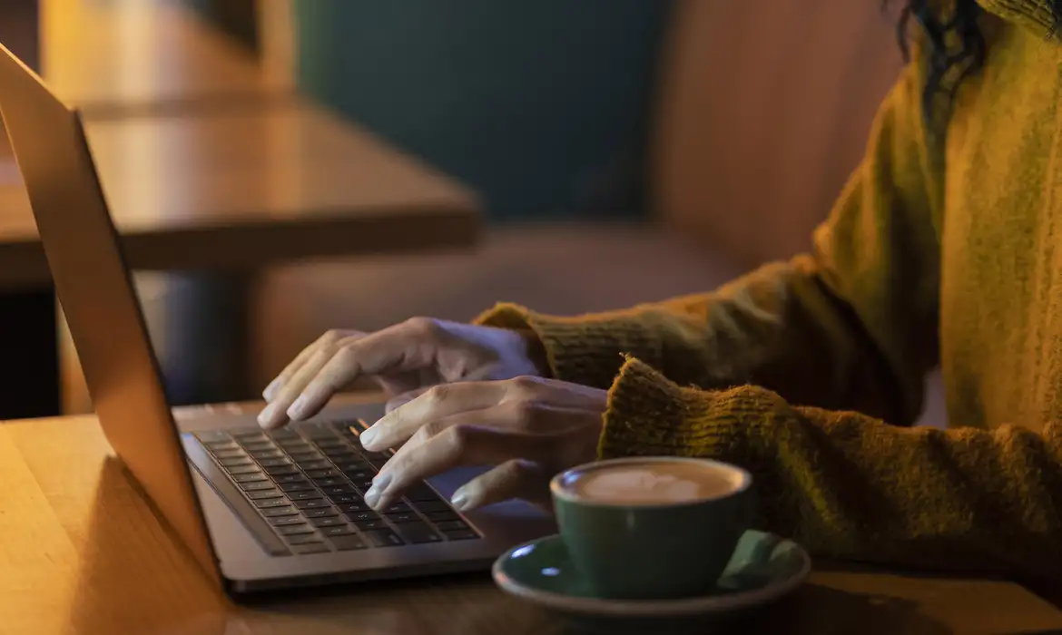 Pessoa digitando no laptop com xícara de café ao lado Mãos femininas digitando no teclado de laptop com xícara de café em mesa de madeira