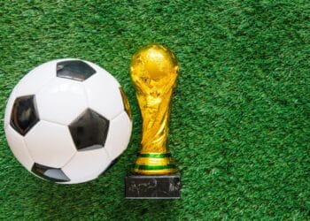 Troféu dourado da Copa do Mundo ao lado de uma bola de futebol em um campo de grama, simbolizando o início do torneio mundial de 2026.