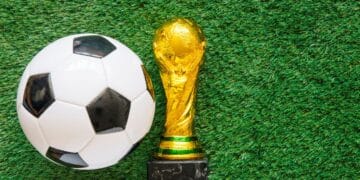 Troféu dourado da Copa do Mundo ao lado de uma bola de futebol em um campo de grama, simbolizando o início do torneio mundial de 2026.