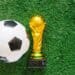 Troféu dourado da Copa do Mundo ao lado de uma bola de futebol em um campo de grama, simbolizando o início do torneio mundial de 2026.