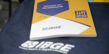 Manual do recenseador IBGE sobre uma bolsa azul com logo do IBGE ao fundo.