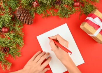 Mãos escrevendo uma mensagem de Natal em papel branco sobre fundo vermelho decorado com ramos e presentes.