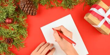 Mãos escrevendo uma mensagem de Natal em papel branco sobre fundo vermelho decorado com ramos e presentes.