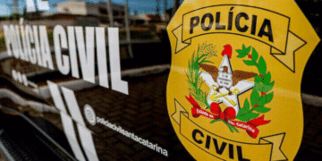 Emblema amarelo da Polícia Civil de Santa Catarina com elementos simbólicos como águia, trigo, café e estrela, afixado em carro policial preto com inscrição "Polícia Civil" ao fundo.