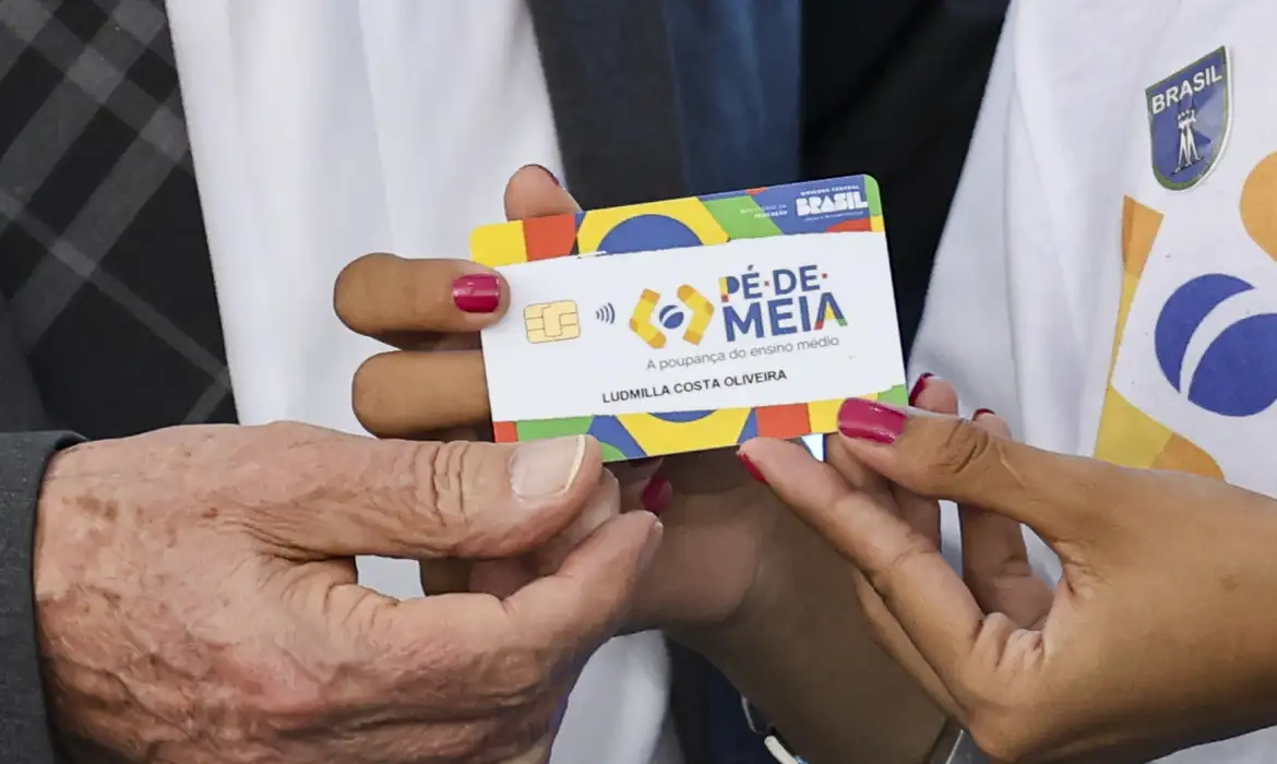 Pé-de-Meia 2026: Como o aumento do salário mínimo pode impactar o programa Imagem do cartão do programa Pé-de-Meia sendo segurado por duas pessoas, simbolizando o auxílio financeiro para estudantes do ensino médio.