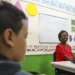 MEC anuncia pagamento de R$ 2.100 por 24 meses: veja quem pode receber o benefício 10 Professora negra sorridente sentada em sala de aula com crianças ao redor