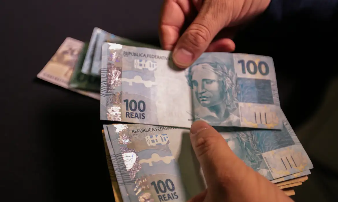 impacto novo salario minimo beneficios sociais 2026 Close das mãos de uma pessoa segurando e contando diversas notas de 50 reais (cédulas de Real), com foco na textura do papel-moeda.