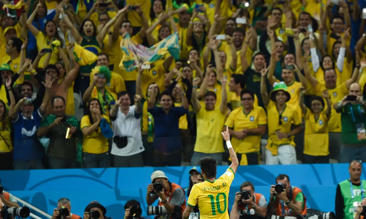 calendario jogos brasil fase de grupos copa 2026 Vista de costas do jogador Neymar Jr. vestindo a camisa 10 amarela da Seleção, com o dedo indicador apontado para o céu; ao fundo, uma arquibancada lotada de torcedores vestindo amarelo celebra com braços erguidos.