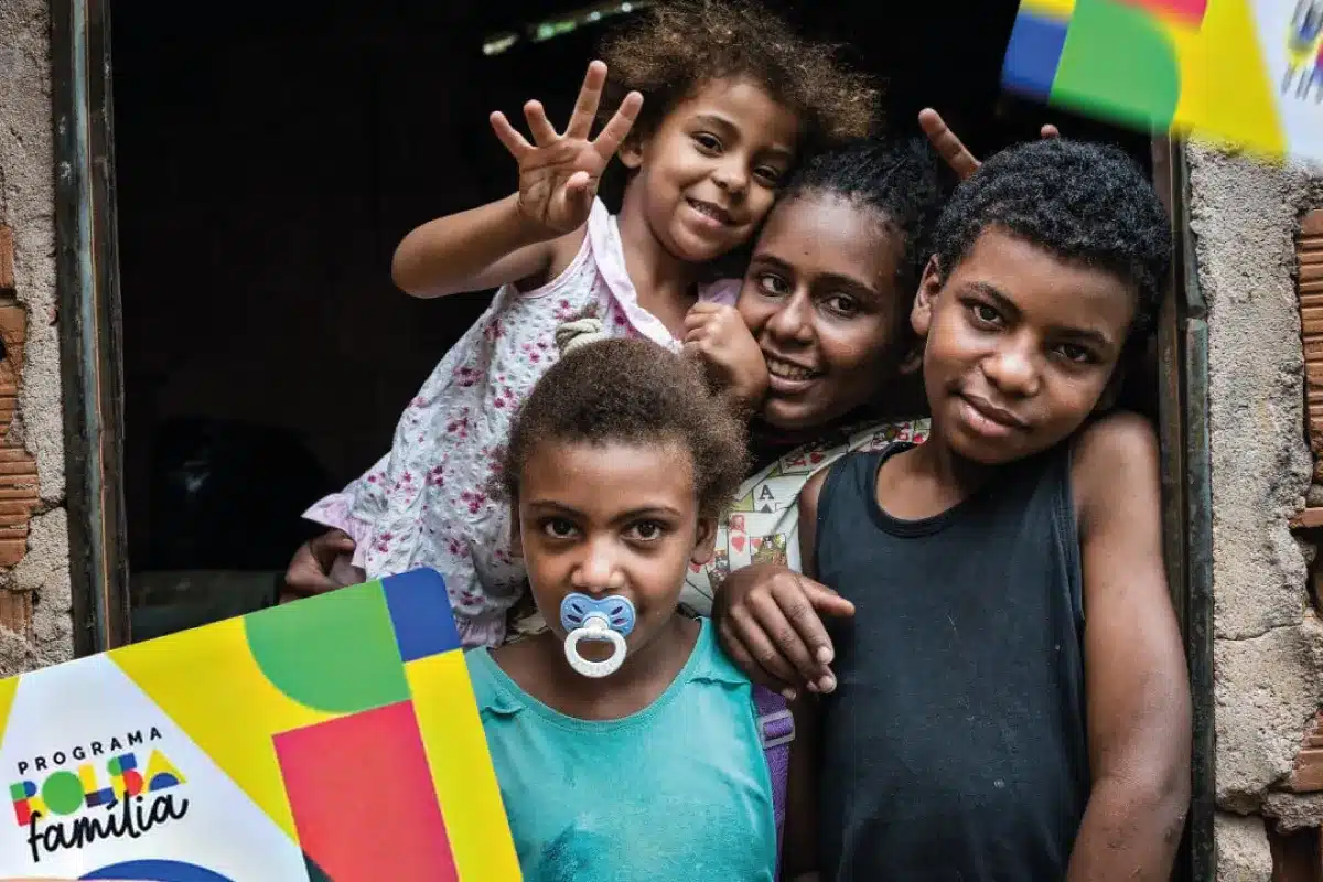 Família beneficiada pelo Bolsa Família em 2026 Quatro crianças sorriem na porta de uma casa simples com cartão do Bolsa Família.
