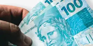 Close-up de uma mão segurando três cédulas de 100 reais chanceladas pelo Banco Central do Brasil, com a efígie da República em destaque sobre o papel-moeda azulado.
