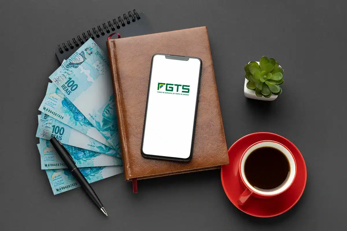 Consulta de saldo FGTS no celular passo a passo Celular mostrando logo do FGTS sobre caderno marrom com cédulas de real, caneta, café e planta