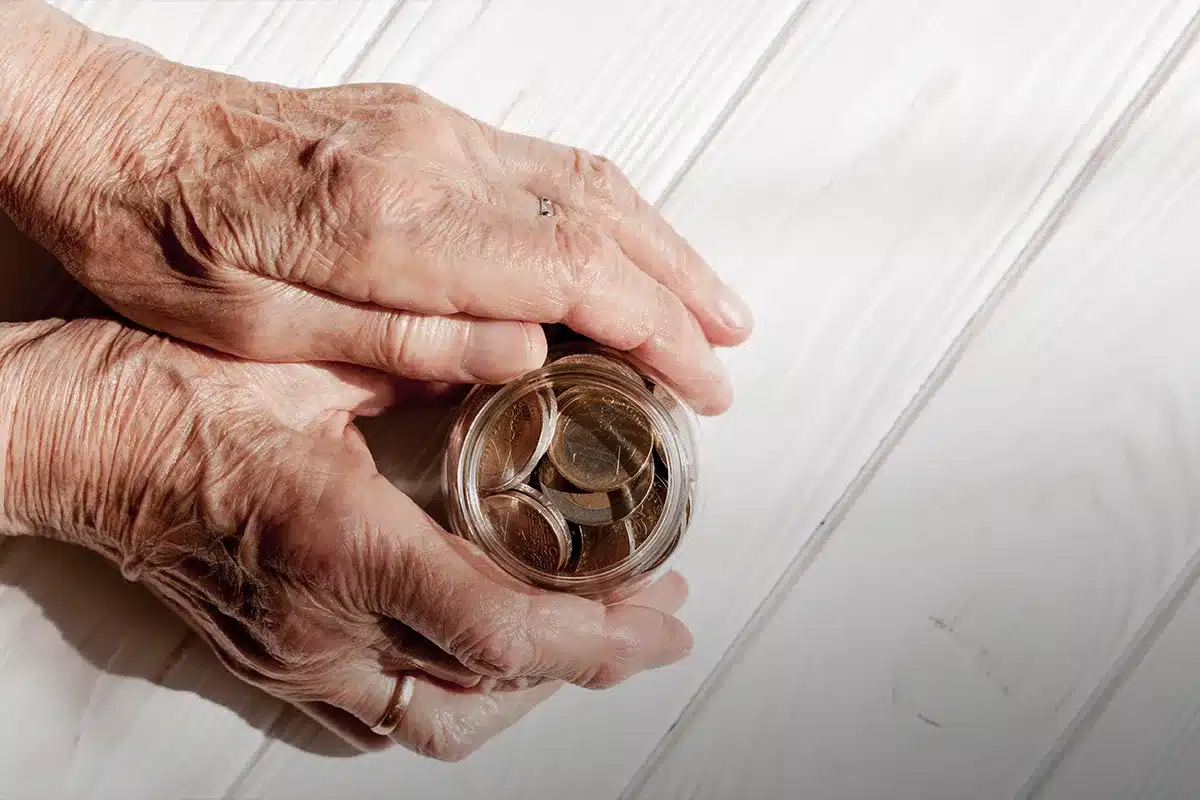 Mãos envelhecidas segurando pote com moedas Mãos de idoso segurando um pote transparente cheio de moedas sobre mesa branca