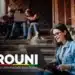 Estudante sorrindo enquanto acessa informações no laptop sobre o Prouni 2026, com outros estudantes ao fundo em uma escada. Inscrições abertas de 26 a 29 de janeiro.